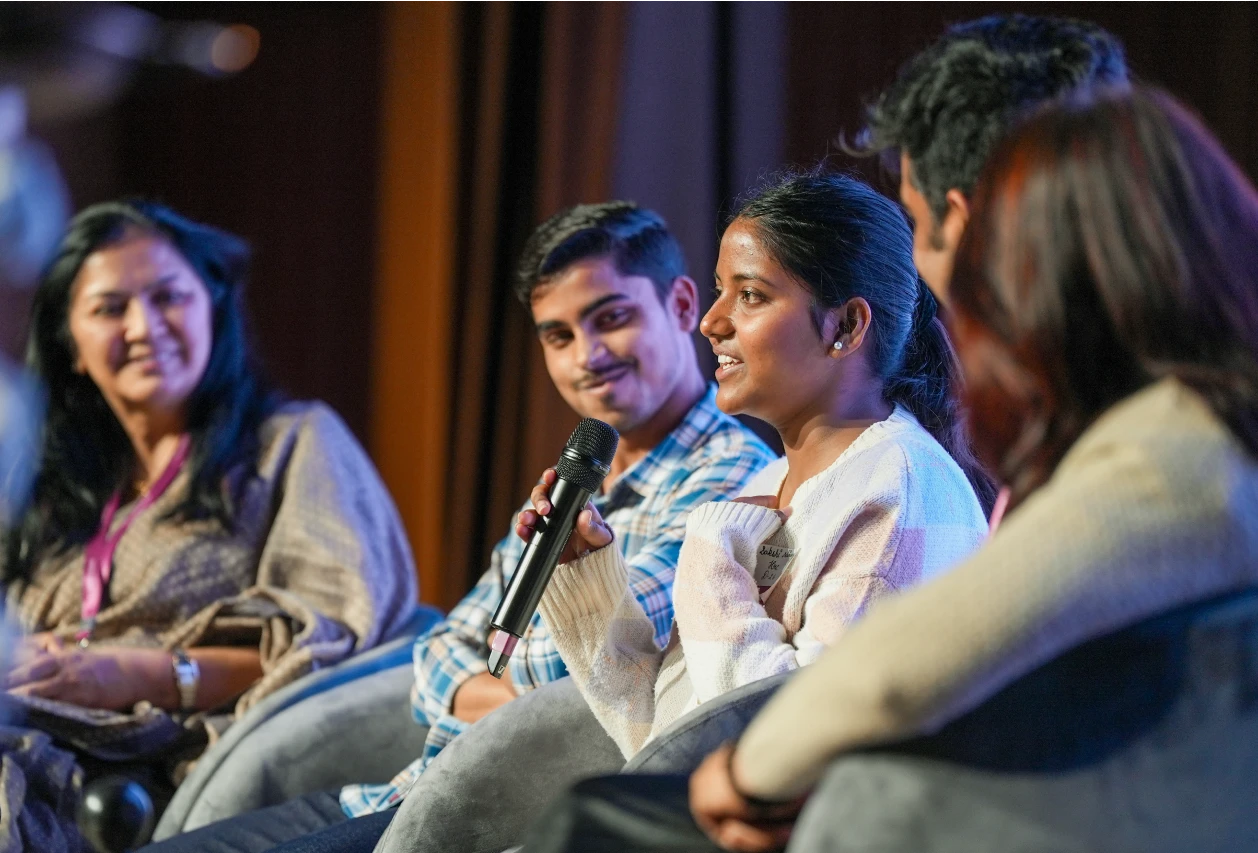 Woman speaking into a microphone at an event, with people listening.