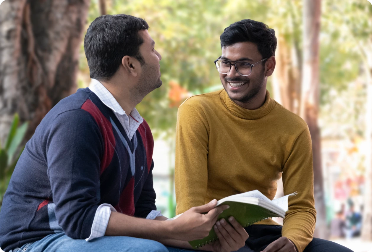 Two men sitting and talking outdoors, one holding a green notebook